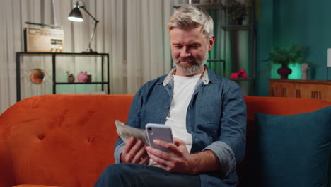 hombre de mediana edad sonriente feliz en casa contando dinero en efectivo uso de teléfono inteligente, ingresos, ahorros, ganar la lotería