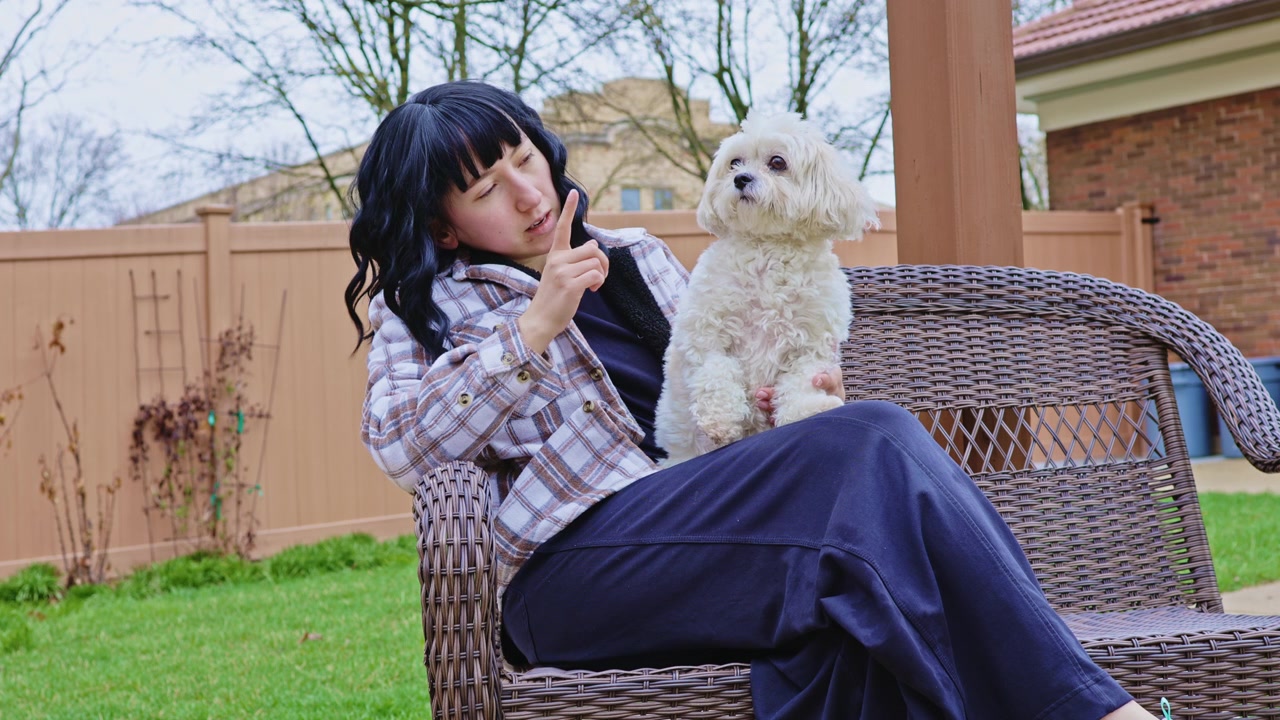 Woman sits on a couch with a small dog in a backyard during a cloudy day while making a gesture with her hand