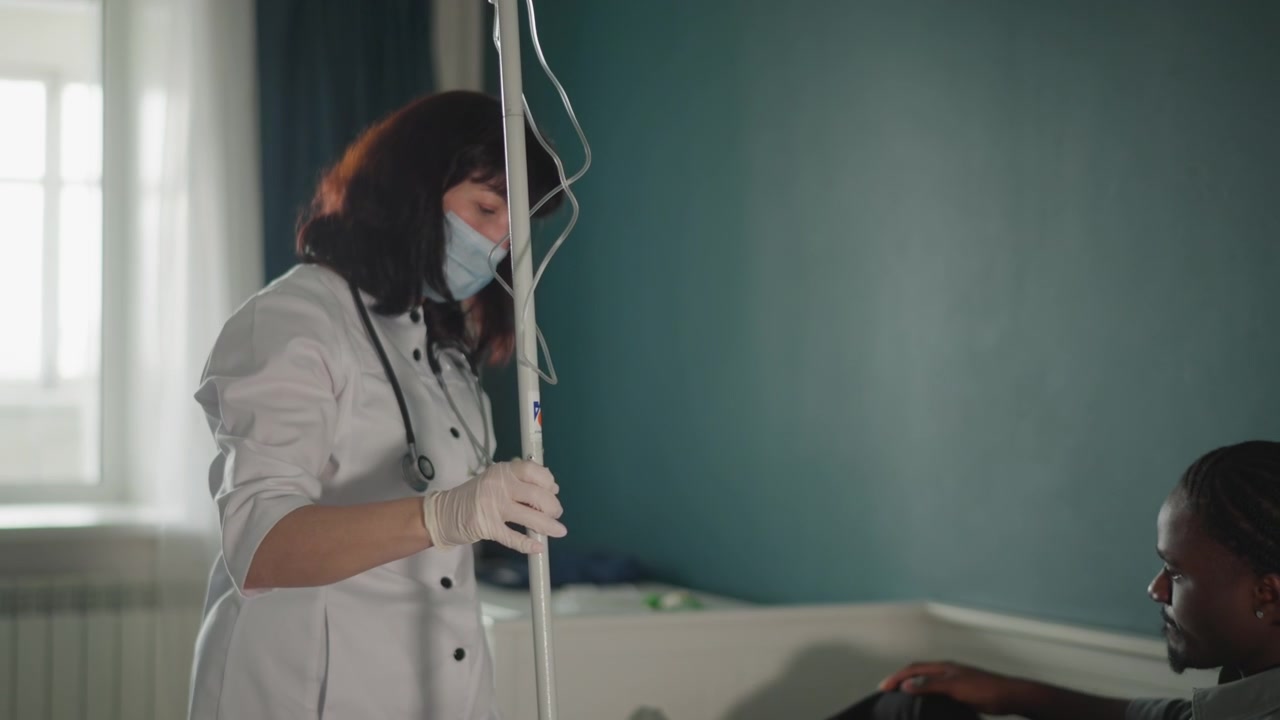Visita medica a domicilio: il medico controlla l'asta della flebo vicino al paziente seduto, il clinico mascherato in camice bianco regola i tubi e monitora il comfort, luce naturale soffusa e un ambiente intimo della camera da letto mostrano una visita a domicilio attenta