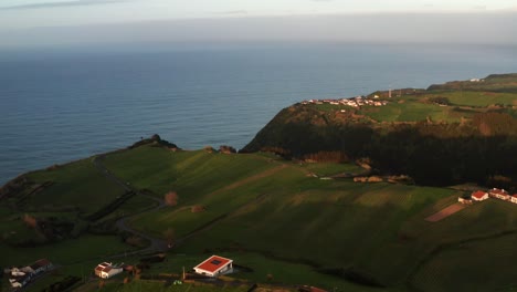 Drone-footage-of-lush-green-volcanic-island-countryside-at-sunset-with-homes-near-ocean-cliff-on-the-Azores-Sao-Miguel-Island