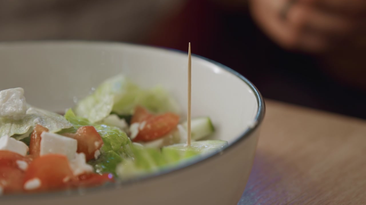 Nærbillede af frisk salat med salat, tomater, agurker og fetaost i en hvid skål. En trætandstikker holder ingredienserne sammen, mens dressing hældes over dem. Baggrunden forbliver blødt sløret.
