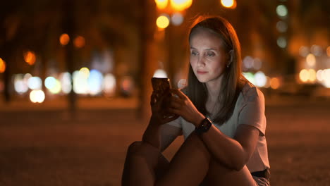 a young woman looks into the smartphone and writes text messages on the internet against the backdrop of the night city. girl businessman working on vacation remote work via mobile phone. gadget addiction