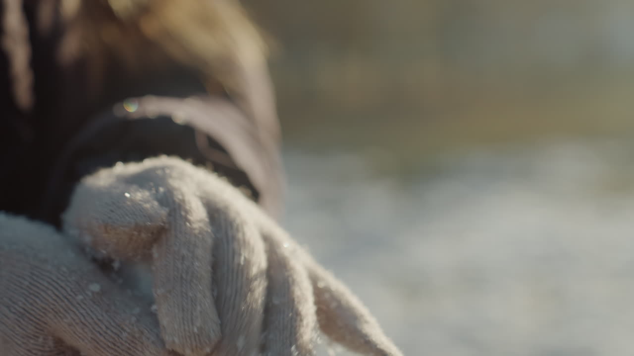Wollhandschuh-Hände formen Schneeball, Sonnenlicht. Nahaufnahme einer Person im Wintermantel, die pulvrigen Schnee zu einer kompakten Kugel formt, sanftes Gegenlicht fängt fliegende Flocken ein, strukturierte Strickfasern mit Schnee bedeckt.