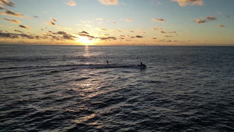 Toma-De-Drone-Que-Muestra-La-Silueta-De-Una-Moto-Acuática-De-Crucero-Llevando-A-Un-Surfista-En-El-Agua-Del-Océano-Durante-La-Puesta-De-Sol-Dorada---Pájaro-Salvaje-Volando-Hacia-La-Cámara