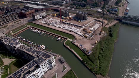 Movimiento-Aéreo-Hacia-Atrás-Que-Muestra-El-Sitio-De-Construcción-De-Un-Edificio-De-Apartamentos-De-Lujo-En-El-Lecho-Del-Río-Ijssel-Que-Revela-El-Paisaje-Urbano-Más-Amplio-Con-El-Horizonte-De-La-Ciudad-De-La-Torre-Zutphen-En-El-Fondo