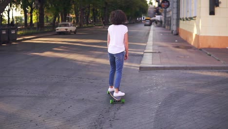 Back-view-of-a-woman-in-a-white-t-shirt,-blue-jeans-and-white-sneakers-skateboarding-in-the-city-street.-Camera-moves-from-legs