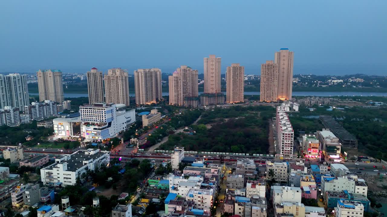 Aerial view captures an urban skyline at dusk with numerous high-rise residential and commercial buildings. A busy illuminated street with traffic runs through the foreground