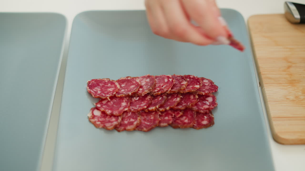 Chef hands placing rows of thin cured sausage slices on sleek tray, highlighting rich red marbling and precise plating technique under soft natural light for elegant appetizer presentation