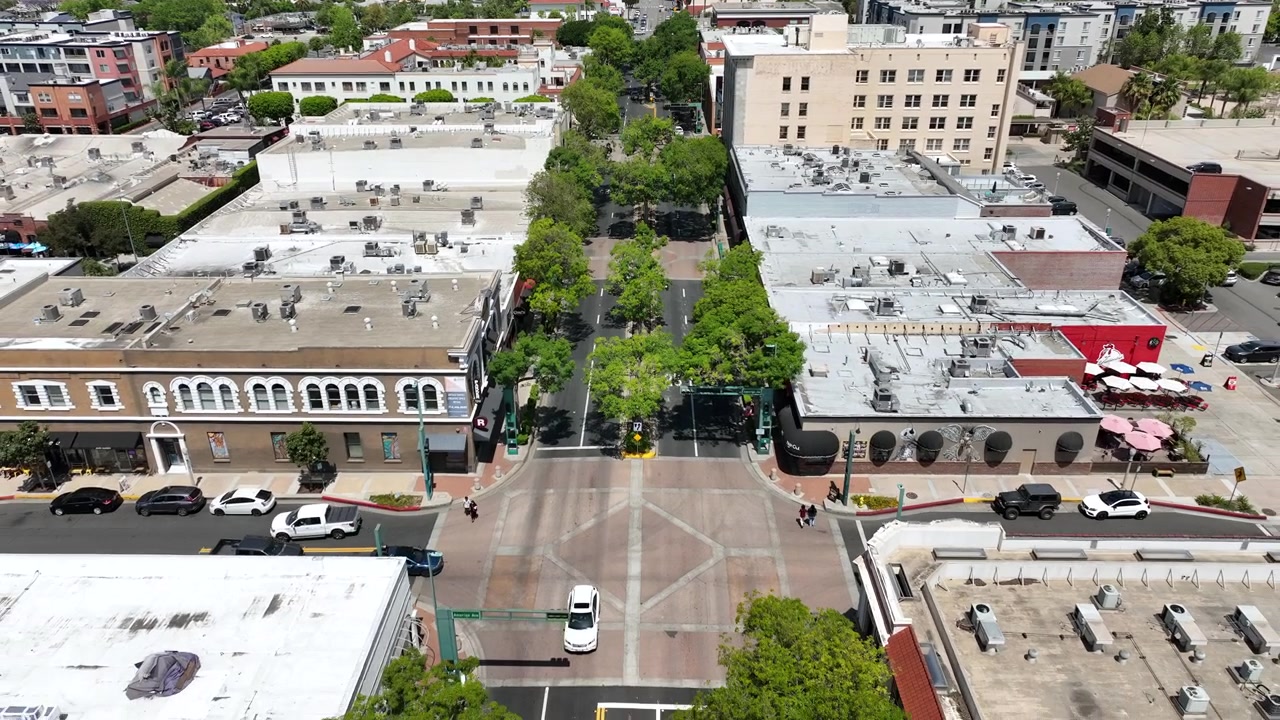 Plan aérien urbain par drone, en recul, de Harbor Boulevard à Fullerton, présentant des bâtiments commerciaux, des terrasses de restaurants, des bars, des touristes et une circulation urbaine active dans le centre-ville.