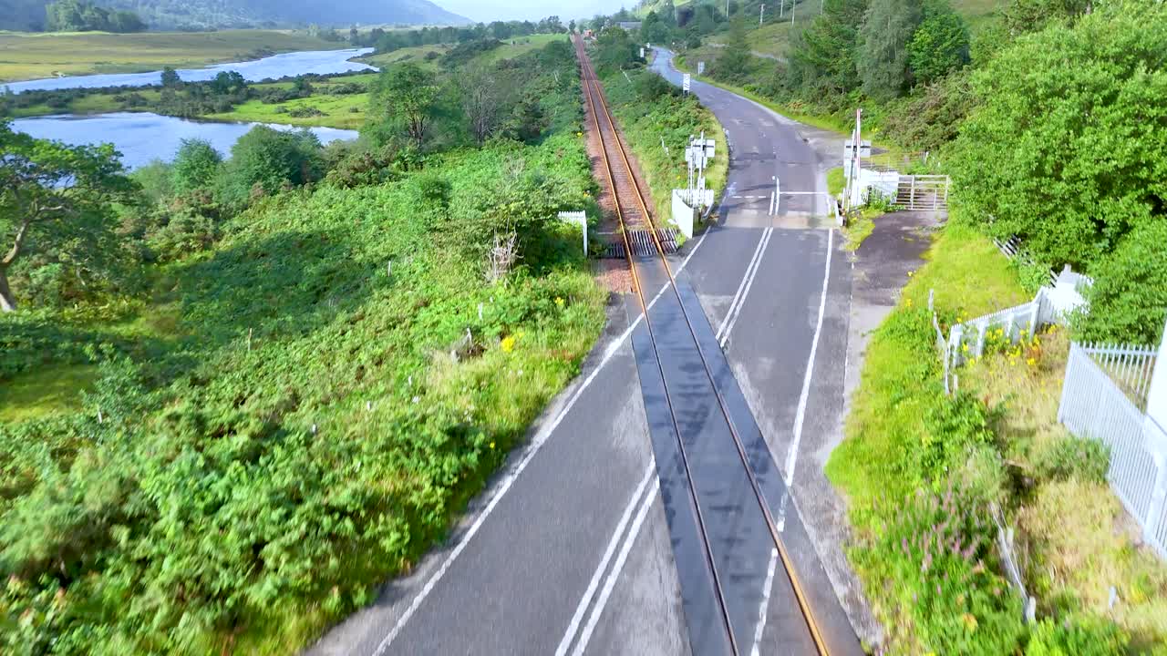 A blue car drives over a railway level crossing in the lush, sunlit Scottish Highlands, captured from an elevated, forward-moving perspective with vibrant greenery and open skies