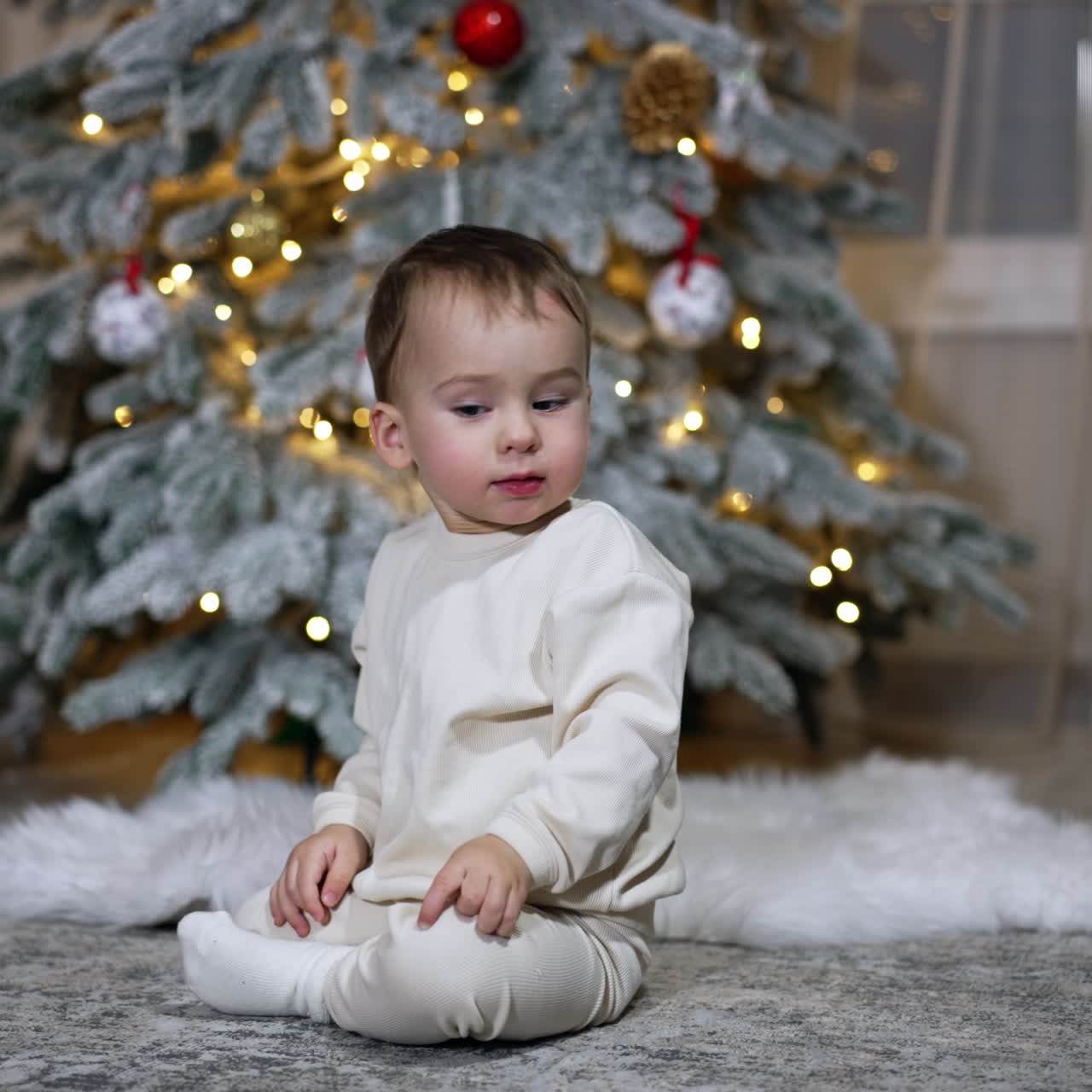 Little lovely boy in white clothes sits on the floor. Baby throws the red ball in front of him. Christmas tree at backdrop