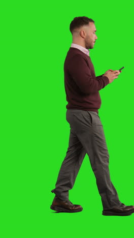 vertical video full length studio shot of businessman using mobile phone to message or browse online walking across green screen