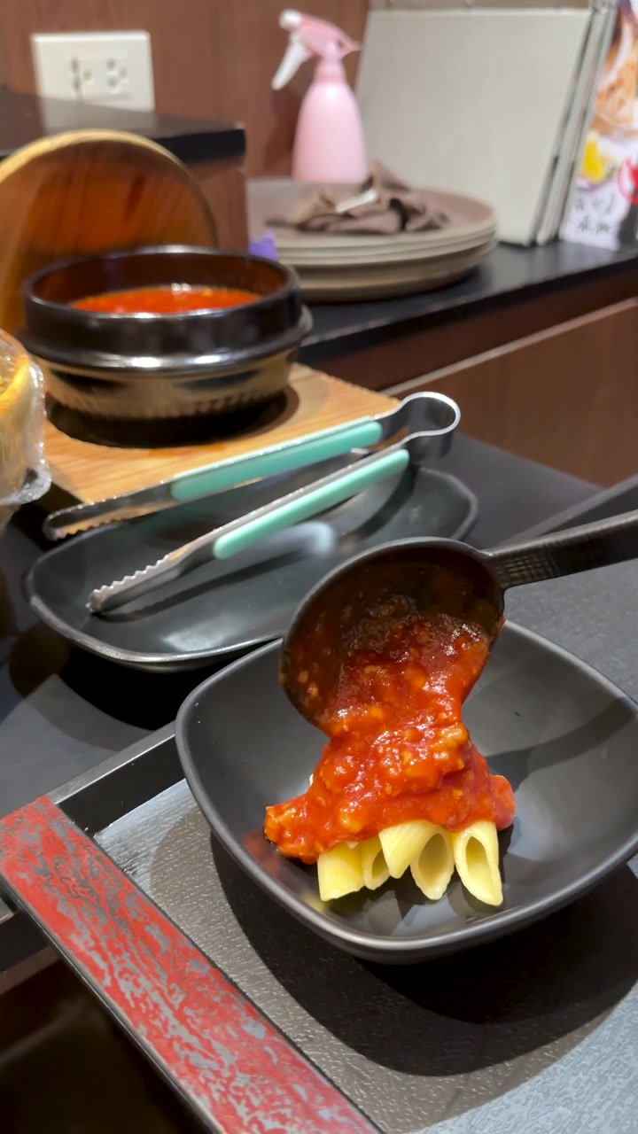 A person ladles red tomato sauce onto penne pasta in a black bowl at buffet.