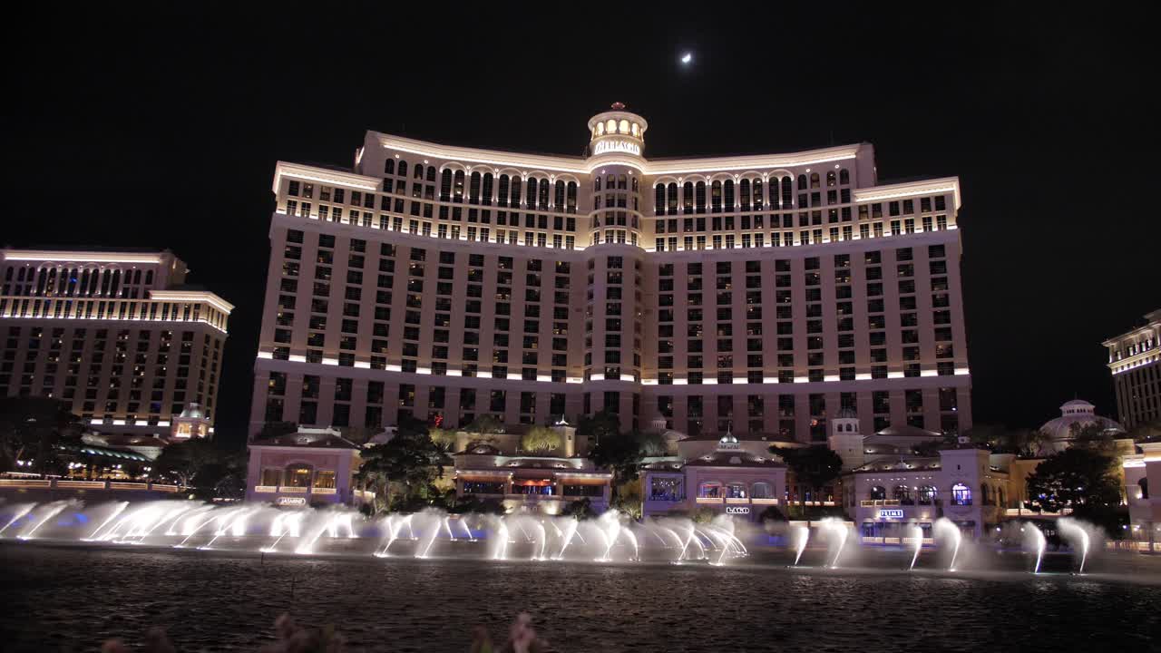 Handheld of luxurious Bellagio Hotel and Casino next to lake with synchronized dancing water fountain illuminated at night, Las Vegas, USA