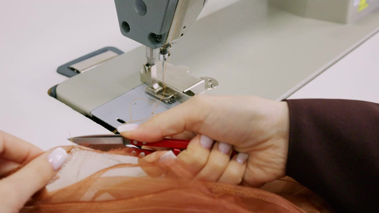 Close up of a designer's hands cutting mustard sewing thread with small red thread snips beside an industrial sewing machine