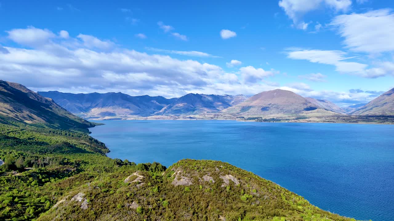 Drone camera smoothly pans across Lake Wakatipu, lush hills, and distant mountains in daylight