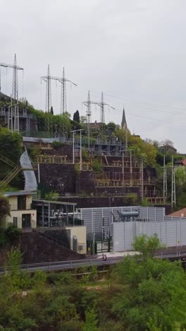 set on a terraced slope in bolzano, the electric station rises beside stairways and pylons, framed by a distant church spire above green trees. vertical drone shot, top-down angle, real time.