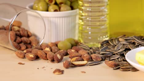 roasted almonds tumble from a tipped glass onto a wooden table with olives, sunflower seeds, and oil, under bright, even lighting and a static camera