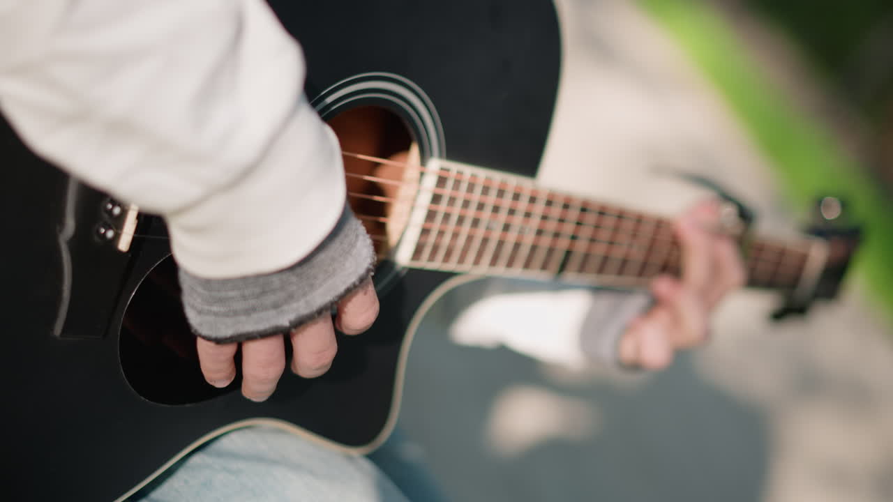 chitarrista di strada vestito di nero che suona una chitarra acustica, performance dinamica da artista di strada sul marciapiede con foro armonico visibile, guanto senza dita e denim, luce solare calda ed effetto bokeh, ritmo allegro e melodia vivace, strada