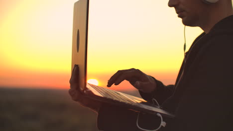 standing at sunset on the roof with a laptop and a beer. a man in a hoodie works having fun and contemplating the beauty of the city view from a height.