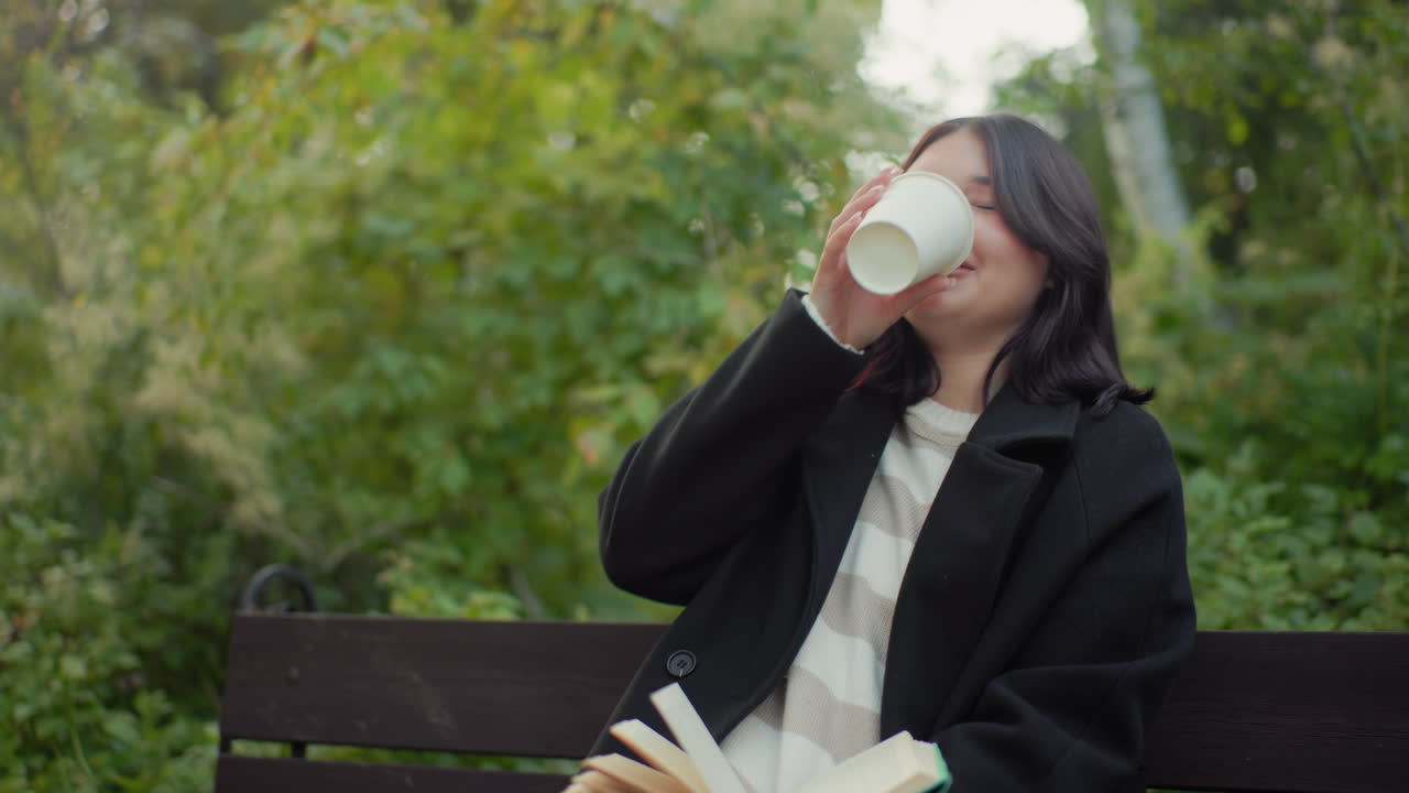 Joyful girl in long jacket sits on park bench, reading novel and flipping pages with soft smile, lifting cup of tea for sip between lines, calm autumn arboretum around with gentle breeze