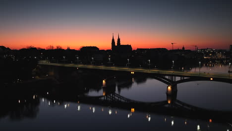 Drohnen-Dolley-Aufnahme-Der-Wettsteinbrücke-Mit-Den-Silhouetten-Des-Münsters-In-Der-Skyline-Der-Stadt-Basel-Nach-Sonnenuntergang