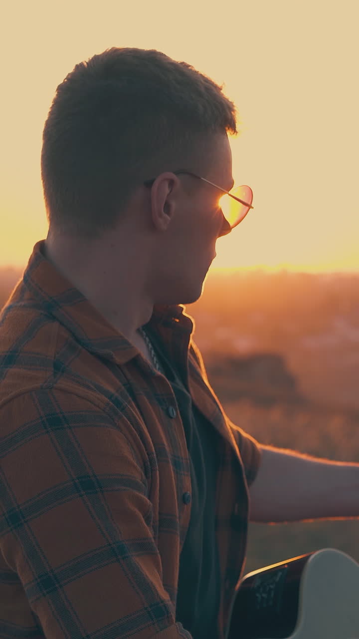 young guy plays guitar in tourist camp against pictorial river and setting sun on horizon in autumn evening close view