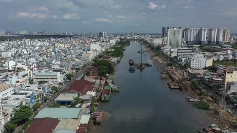 Drone-flight-along-Kenh-Te-canal-in-Ho-Chi-Minh-City-Vietnam-on-sunny-afternoon-with-river-boats,-waterfront-houses,high-rise-buildings-and-reflections