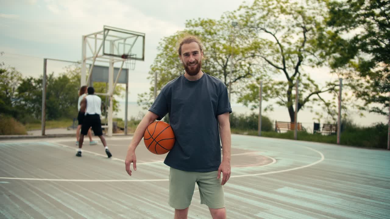 un hombre pelirrojo en una camiseta gris hecha de un baloncesto naranja posa contra el fondo de sus amigos en la cancha de baloncesto afuera en el verano