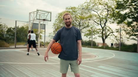 a red-haired man in a gray t-shirt made of an orange basketball poses against the background of his friends on the basketball court outside in the summer