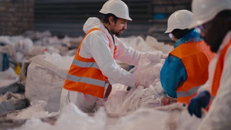 A-brunette-man-with-a-beard-in-a-white-protective-uniform-and-an-orange-vest-presses-cellophane-and-polyethylene-together-with-his-colleagues-at-a-large-waste-recycling-plant