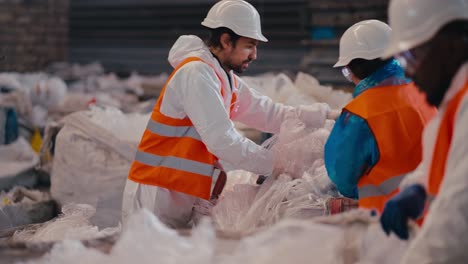 un hombre moreno con barba en un uniforme de protección blanco y un chaleco naranja presiona celofán y polietileno junto con sus colegas en una gran planta de reciclaje de residuos
