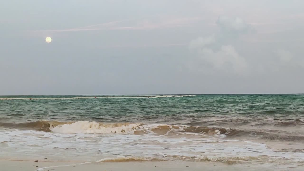Waves are crashing on the beach on an early evening with the full moon out