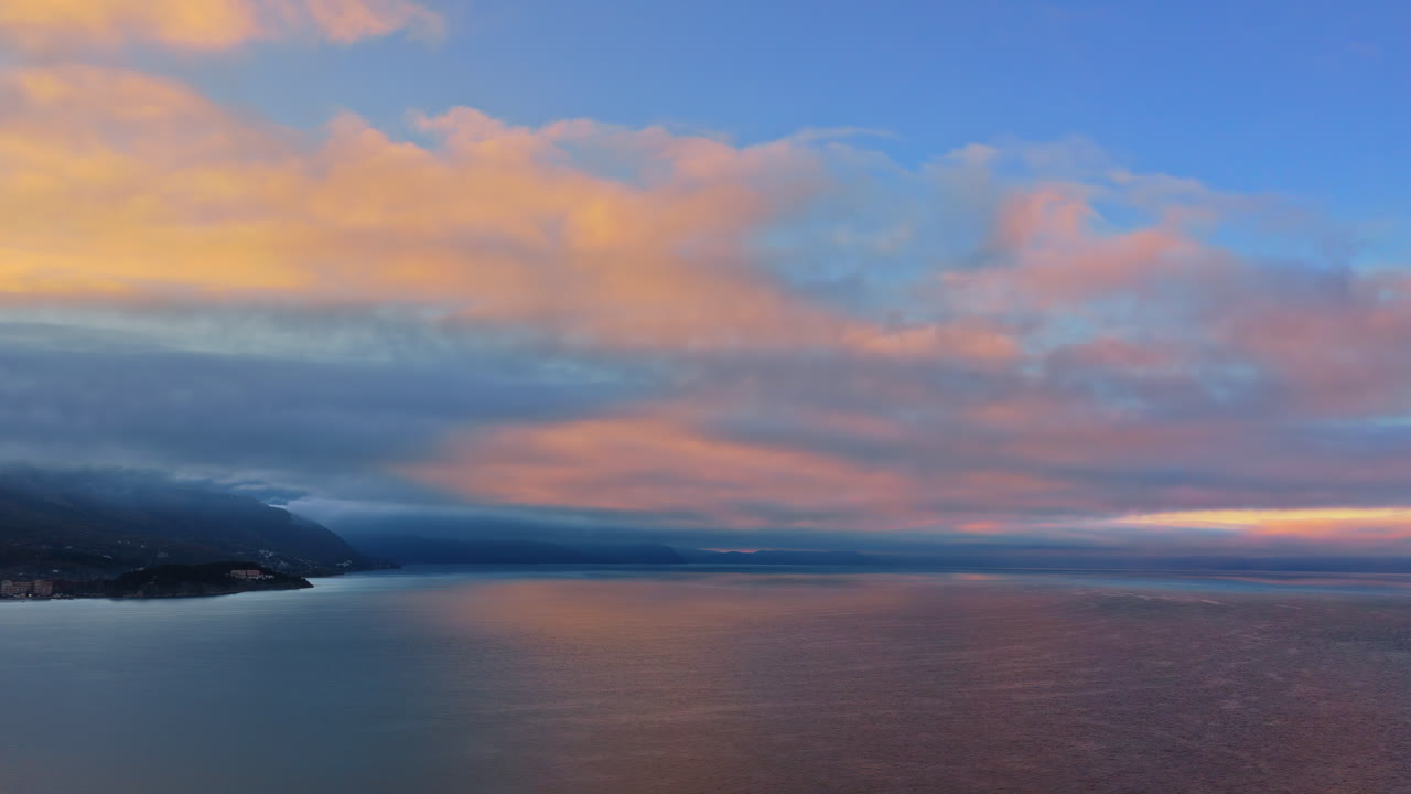 Aerial drone view of Lake Ohrid, with low clouds hugging the mountains and soft pastel light reflecting across the water. North Macedonia