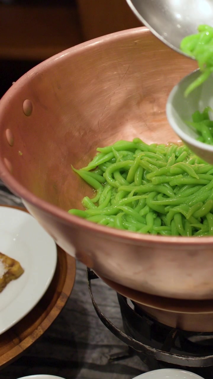 A metal ladle scoops green pandan noodles from a copper pot in a restaurant setting