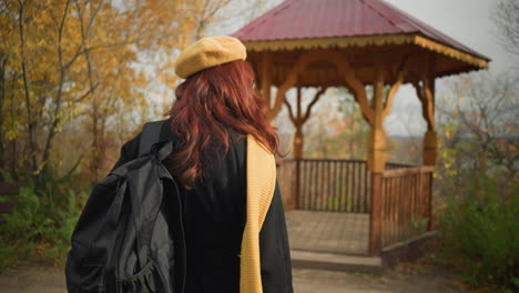 back view of elegant lady in black coat, yellow scarf, and beret walking into wooden gazebo in serene autumn park, wooden bench nearby enhances peaceful atmosphere surrounded by golden trees