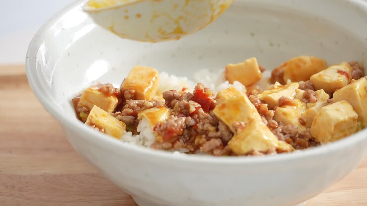 Pouring stir-fried mapo tofu with hot spicy sauce over white rice in a bowl at home