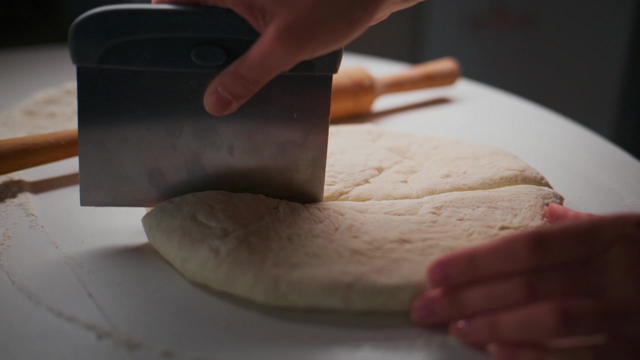 Hand cuts pizza dough with a metal cutter on a white surface. Another hand holds the dough steady. Wooden roller lies nearby, unused. The scene focuses on food preparation