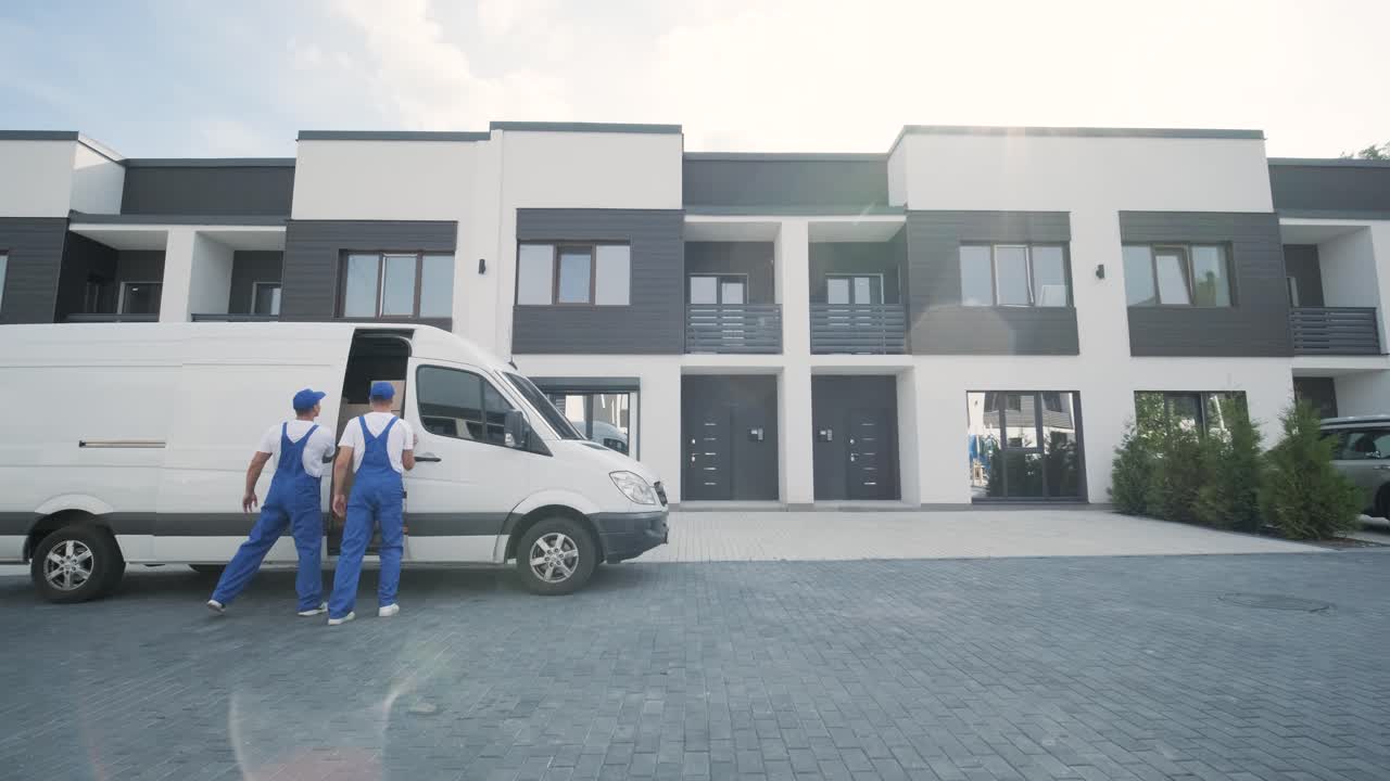 Two young workers of removal company unload boxes and furniture from minibus into customer's home