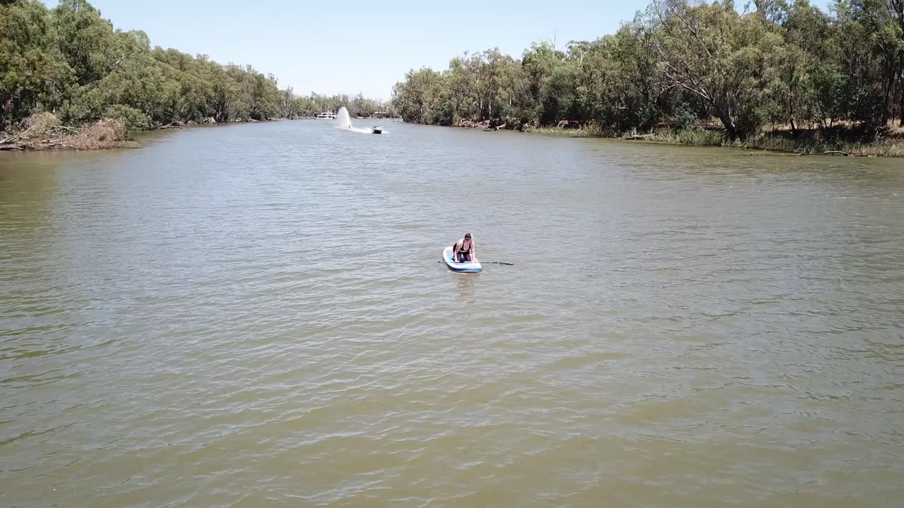 dron aéreo sobre el río mujer arrodillada en stand up paddle board pasa con esquiador acuático