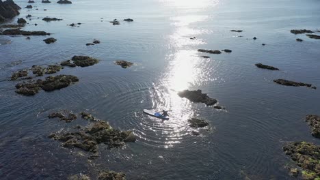 Aerial-view-of-climbing-onto-Stand-Up-Paddle-board-on-calm-water-with-sun-reflecting-along-the-rocky-coastline-in-the-South-of-Ireland-on-a-calm-day