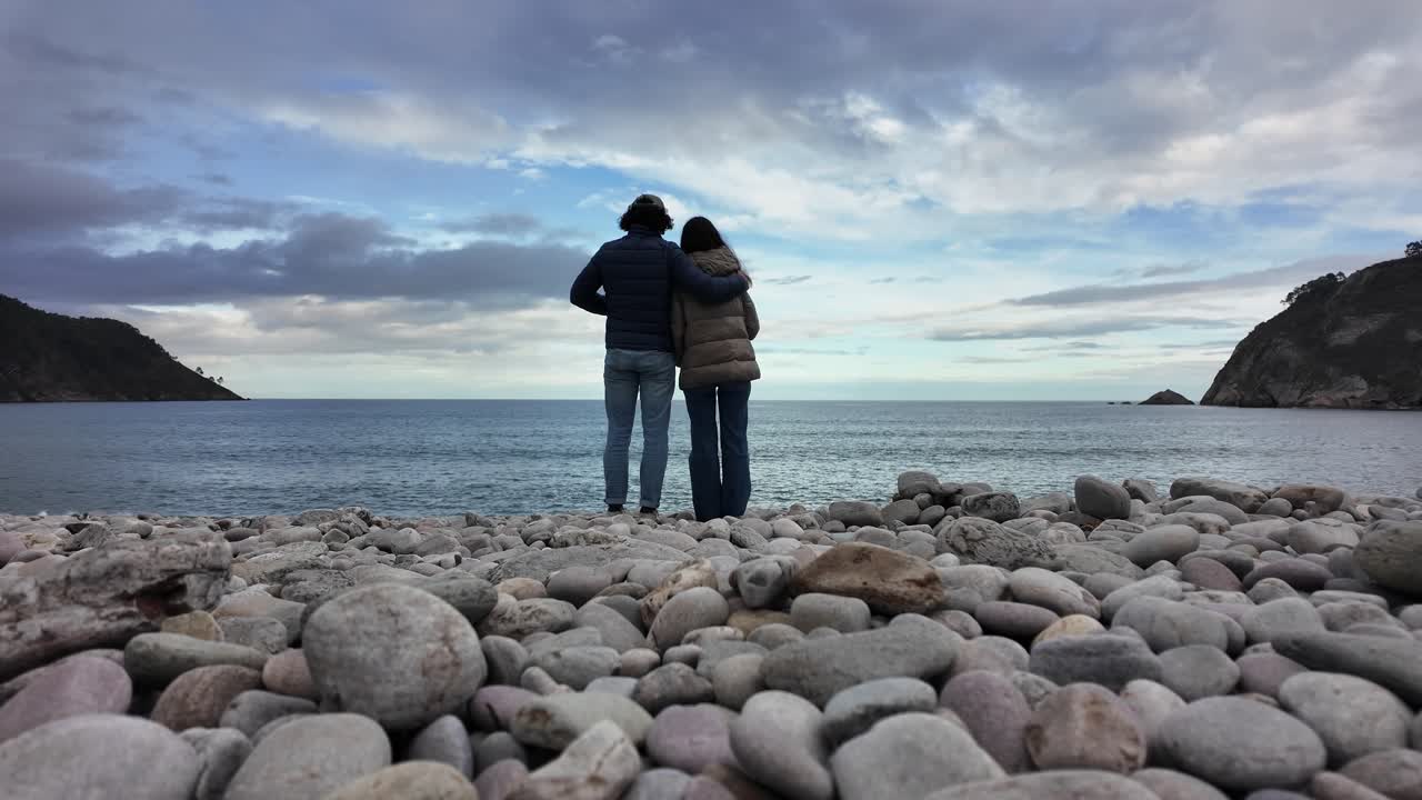 Two people stand close together on a pebble beach, watching the calm sea beneath cloudy skies. Love