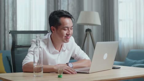 아시아인 남자가 집에서 노트북으로 비디오 통화를 하는 동안 유리잔에서 보드카를 마시고 있다