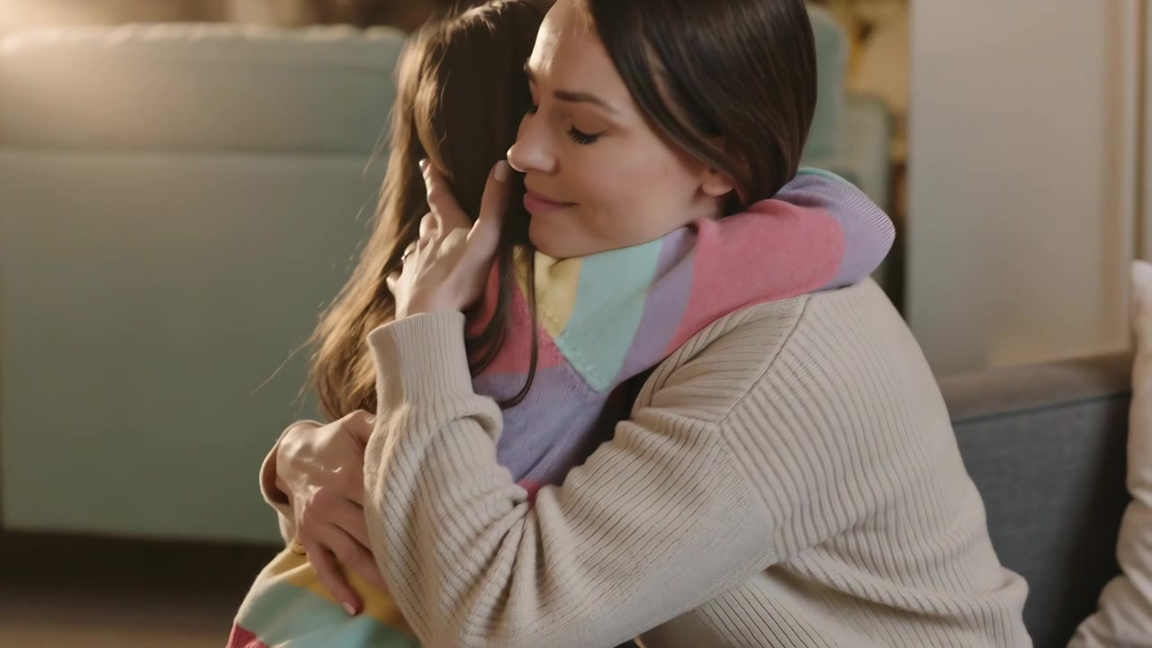 Madre e figlia che si abbracciano sul divano, con la bambina in maglione arcobaleno che inizia l'abbraccio per conforto.