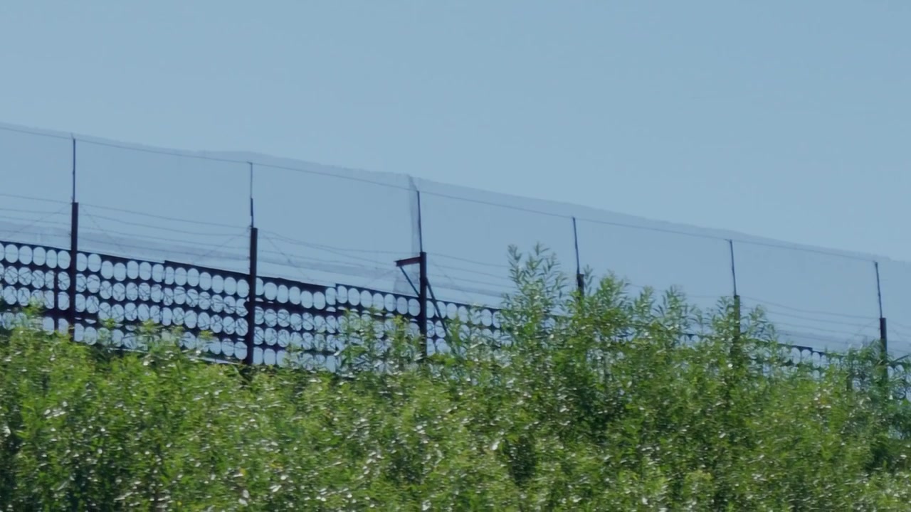 Close up tracking shot shows design of high metal border security fence with circular patterns and barbed wire on top of hill behind green foliage in Latvia.