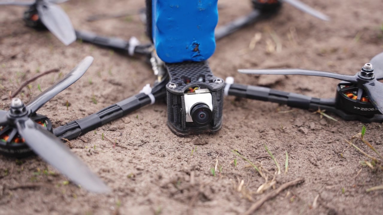Yksityiskohtainen lähikuva FPV-dronen komponenteista likaisella maalla, esittäen kameran ja rungon asettelua.