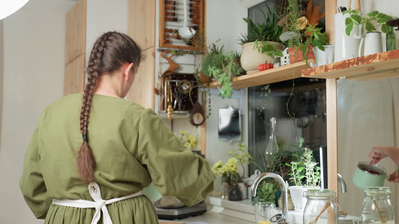 Back view of woman with long braid wearing green gown preparing breakfast as she reaches to operate electric mixer in cozy kitchen with wooden shelves