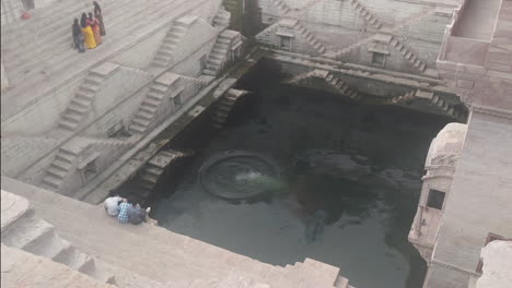 Tres-Niños-Miran-Mientras-Un-Hombre-Con-Pantalones-Cortos-Negros-Se-Sumerge-En-Un-Hermoso-Pozo-En-Jodhpur,-India,-Conocido-Como-Toorji-Ka-Jhalra-O-Pozo-De-Paso-De-Toorji.