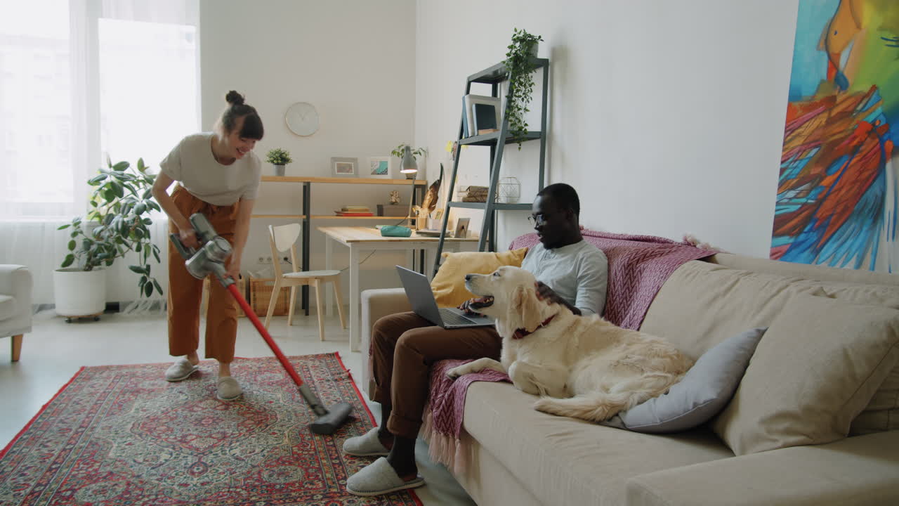mujer aspirando el piso mientras el marido y el perro sentados en el sofá