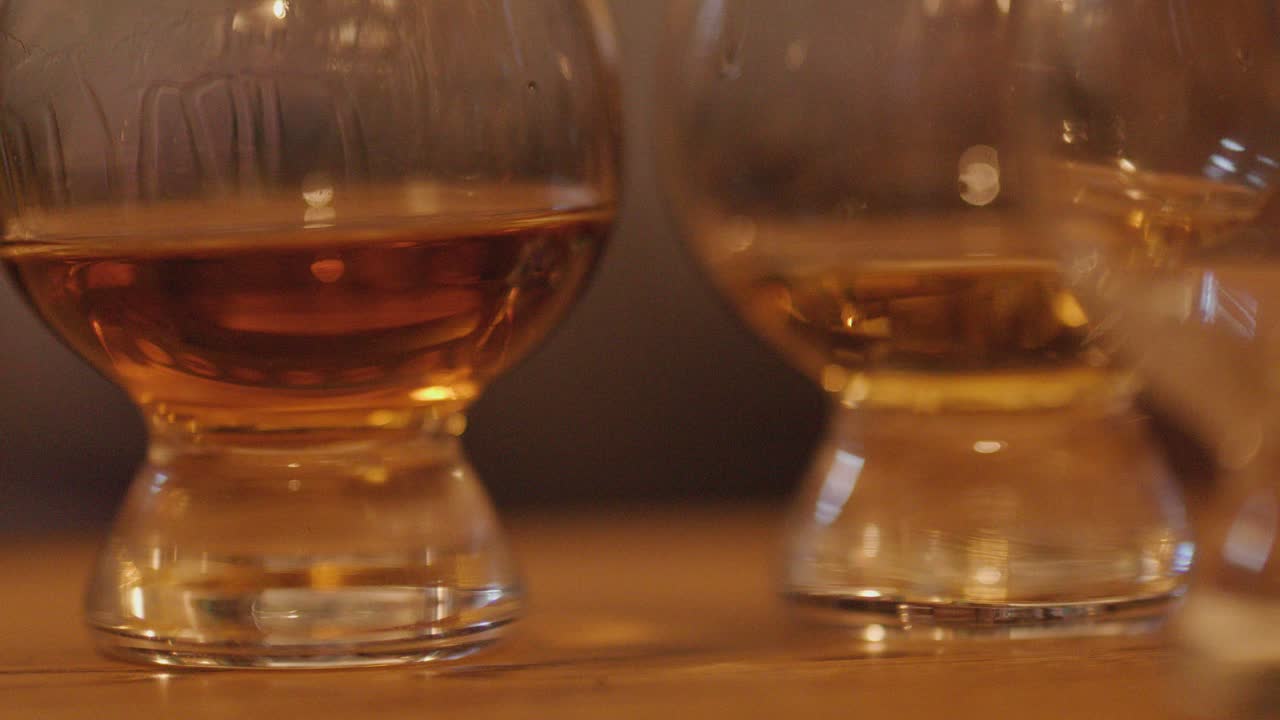 Close-up of whiskey being gently swirled in a glass beside another, with warm ambient lighting and shallow depth of field on a wooden bar
